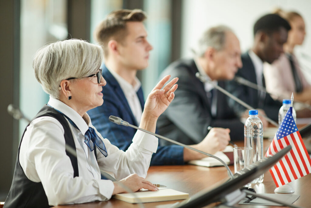 Senior woman with gray hair and glasses speaking at a conference table with a microphone in front and American flags on the desk.