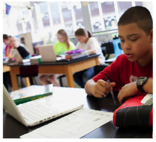 Student in a red shirt writes on a worksheet at a desk in a classroom; a white laptop sits nearby as other students work at tables in the background