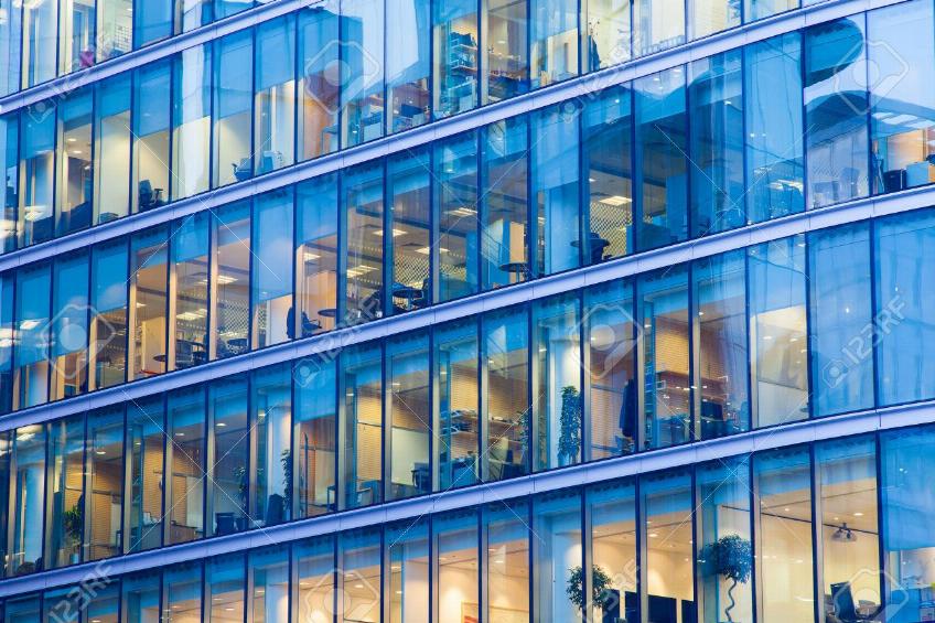 Modern glass-office building with illuminated workspaces visible through blue-tinted windows in diagonal façade view.