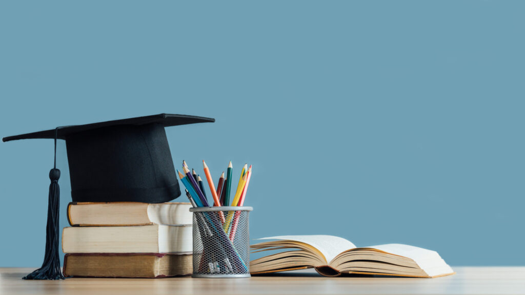 Graduation cap on a stack of books beside a pencil cup of colored pencils and an open book on a desk.
