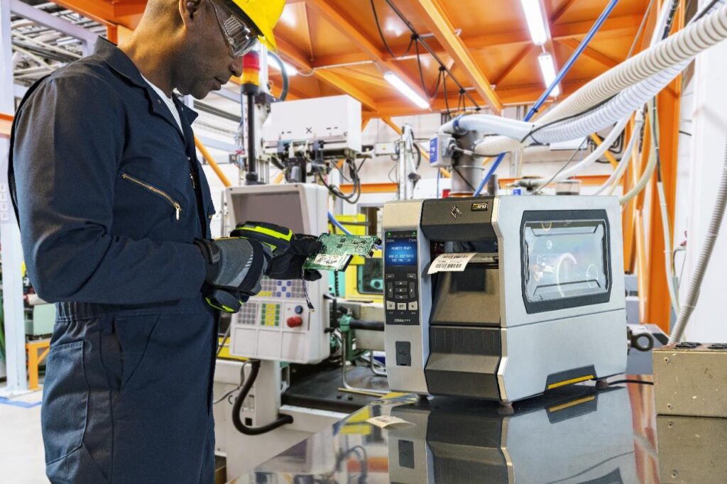 Industrial worker in a yellow hard hat, safety goggles, and dark coveralls inspects a green circuit board next to a large stamping/printing machine in a bright factory setting.