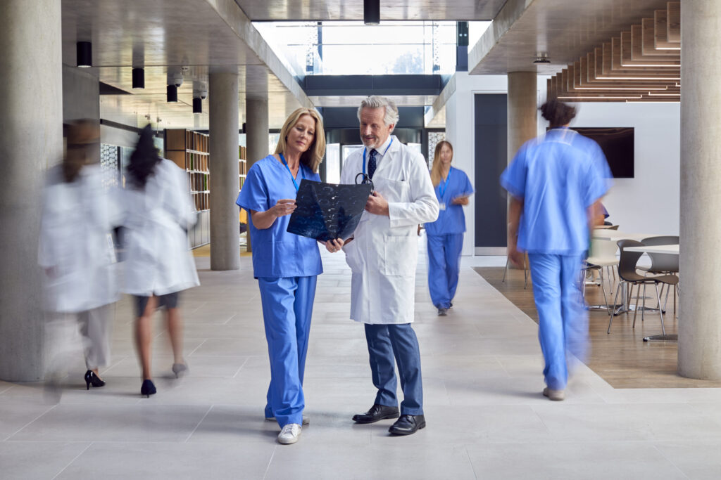 Medical professionals review a patient scan in a bright hospital hallway, with a senior doctor in white coat and a nurse in blue scrubs examining a film