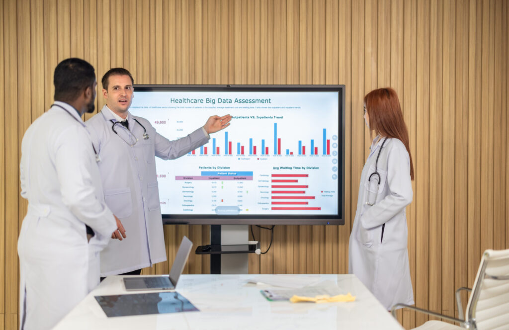 Three doctors in white coats discuss a large screen displaying a data chart titled 'Healthcare Big Data Assessment' in a conference room.