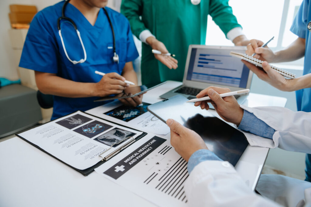 Medical team reviews charts and documents on a table, with a tablet, laptop, and stethoscope in view during a discussion.