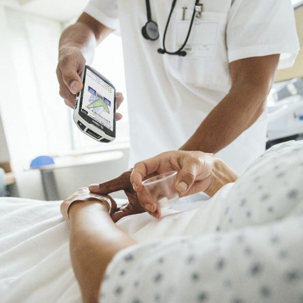 Healthcare professional in white scrubs uses a handheld device to check a patient in bed, stethoscope around neck