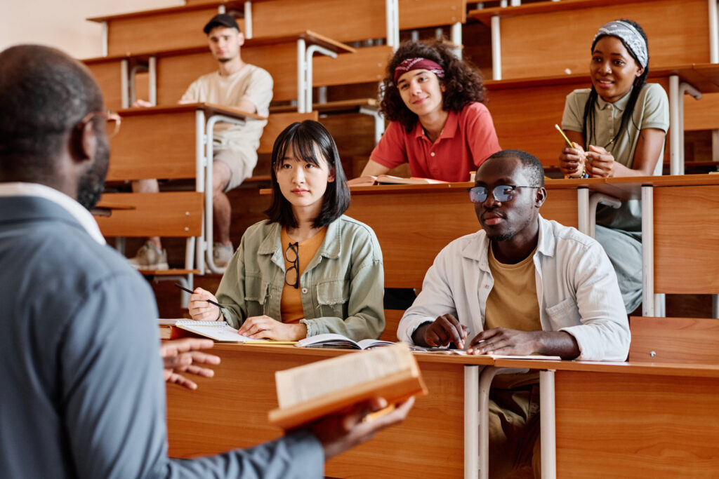Professor addressing a class in a lecture hall as students listen and take notes at wooden tiered desks