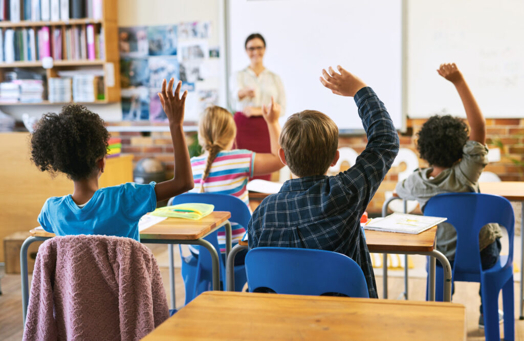 Classroom scene with students raising hands as a teacher walks to the whiteboard at the front.