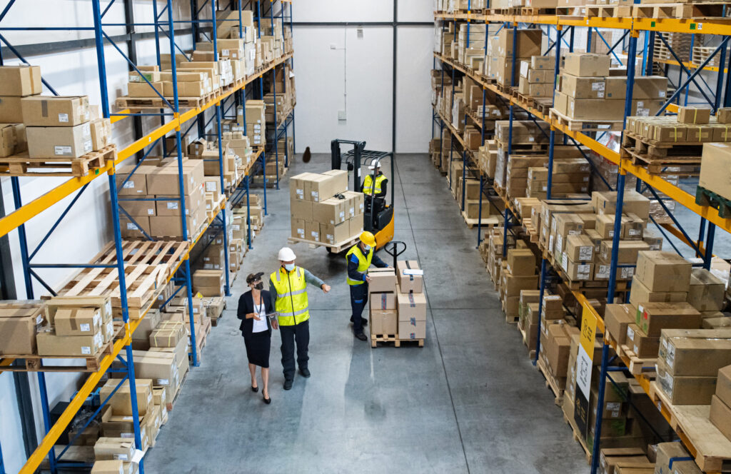 Warehouse with tall blue-and-orange racks full of cardboard boxes; workers in safety vests handling shipments on a concrete floor.