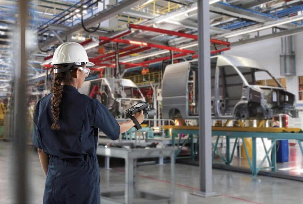 Female factory worker in a white hard hat and safety goggles uses a handheld scanner on an automotive assembly line.
