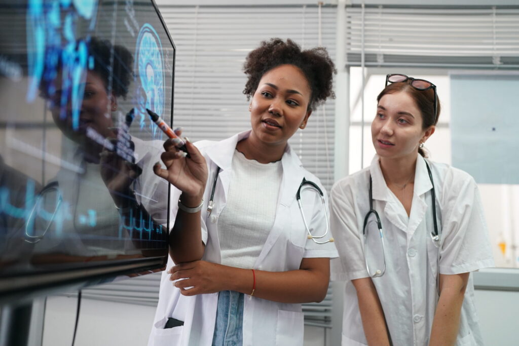 Two female doctors in white coats study a glass touchscreen with blue medical data, one pointing with a marker.
